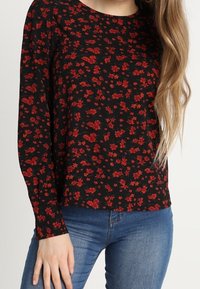 Haut noir à manches longues avec un motif floral rouge, col rond et coupe décontractée. Associé à un jean bleu pour un look casual.
