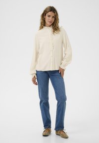 Femme debout portant un chemisier à volants crème, un jean bleu et des ballerines beiges avec des embellissements, sur un fond blanc uni.