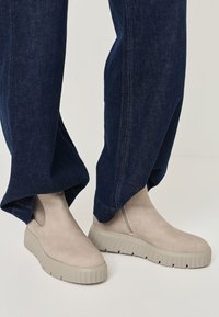 Beige suede ankle boots with textured gray chunky soles, worn with dark blue wide-leg jeans. Notable stitching and elastic side details.