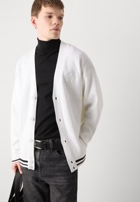 Cárdigan ligero blanco con un patrón texturizado, que cuenta con un cierre de botones, acentos negros en los puños y un cuello de tortuga negro superpuesto.