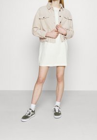 Osoba v bílých šatech, světle béžové zkrácené bundě, bílých ponožkách a zelených teniskách stojící na jednobarevném světlem pozadí.