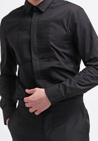 Musta pitkähihainen paita, jossa on teksturoitu pystyraita edessä, nappikaulus ja sileä kangas. Istuva malli.