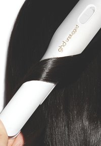 Un lisseur à cheveux ghd débranché blanc pinçant une section de longs cheveux sombres et brillants sur un fond blanc.