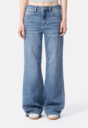 Person iført løse, vidde blå jeans og hvide sneakers, stående mod en ensfarvet lys baggrund.
