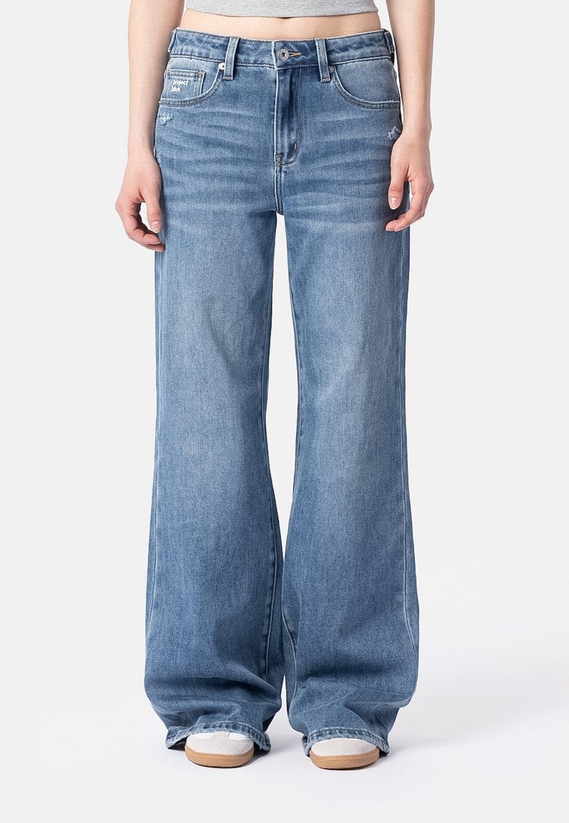 Personne portant un jean bleu large et ample et des baskets blanches, debout sur un fond clair uni.