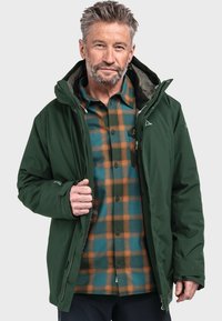 Groene waterdichte jas met een capuchon, voorzien van meerdere zakken, gedragen over een blauw en oranje geruit overhemd. Dikke, gestructureerde stof.