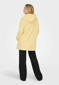 Svetlo rumena dežna jakna s kapuco, sproščenim krojem in dolgimi rokavi, združena s črnimi širokimi hlačami. Gladek tekstura, preprost dizajn.