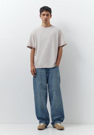 Jonge man staat tegen een witte muur, draagt een oversized beige t-shirt, ruime blauwe jeans en beige sneakers, met één hand in zijn zak.