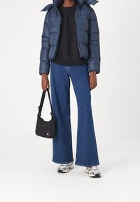 Personne portant une doudoune bleu marine, un pull noir, un jean évasé bleu, des baskets blanches et portant un petit sac à bandoulière noir sur fond blanc.