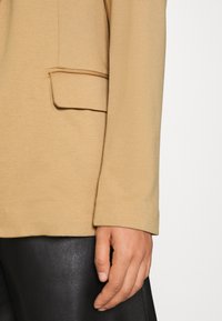 Manche de blazer beige avec détail de poche à rabat sur pantalon en cuir noir, montrant une main détendue reposant à côté de la cuisse.