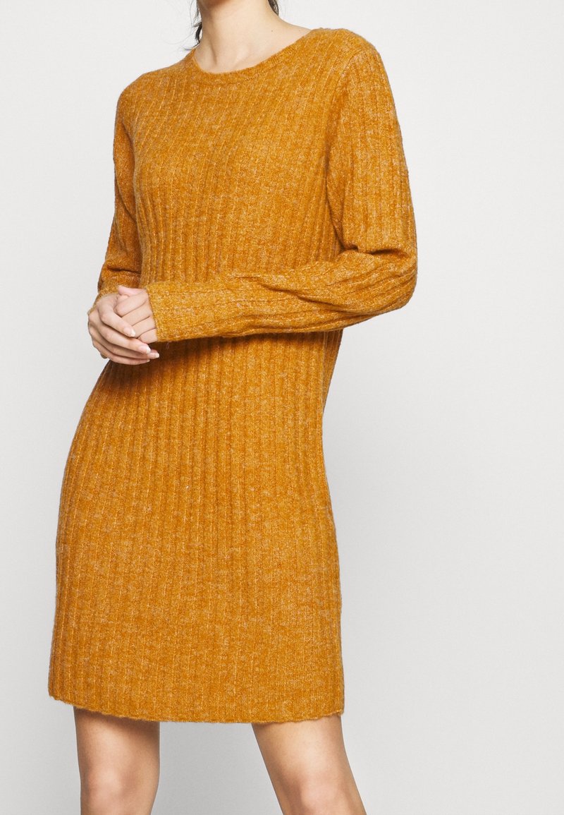 Femme portant une robe-pull à manches longues en maille côtelée de couleur jaune moutarde, debout les mains jointes au niveau du ventre devant un fond uni.