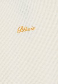 Tejido de ribete crema con líneas texturizadas. Presenta el nombre bordado "Blhois" en hilo dorado en contraste.