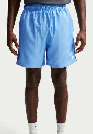 Homme portant un short de sport Nike bleu clair avec une taille élastique et un cordon, associé à des chaussettes blanches Nike, debout devant un fond uni.