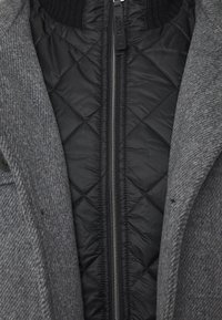 Primer plano de una chaqueta negra acolchada con cremallera, superpuesta bajo un abrigo gris de lana texturizada con un botón visible.