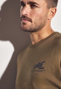 Olivově zelené bavlněné tričko s krátkým rukávem. Na přední straně je modrý textový logo "Custom Made" s dalším textem pod ním. Jednoduchý kulatý výstřih.