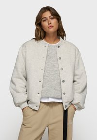 Veste sweat gris clair avec un col arrondi, des boutons-pression et des épaules tombantes. Portée par-dessus une chemise grise et un pantalon beige.