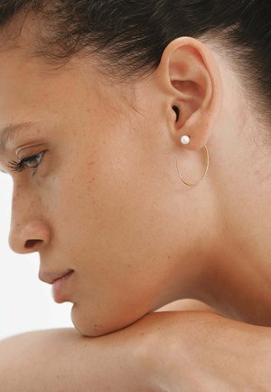 Pendiente de aro de oro con un detalle de perla, colocado en el lóbulo de la oreja, mostrado sobre piel suave con una textura ligera y tonos naturales.
