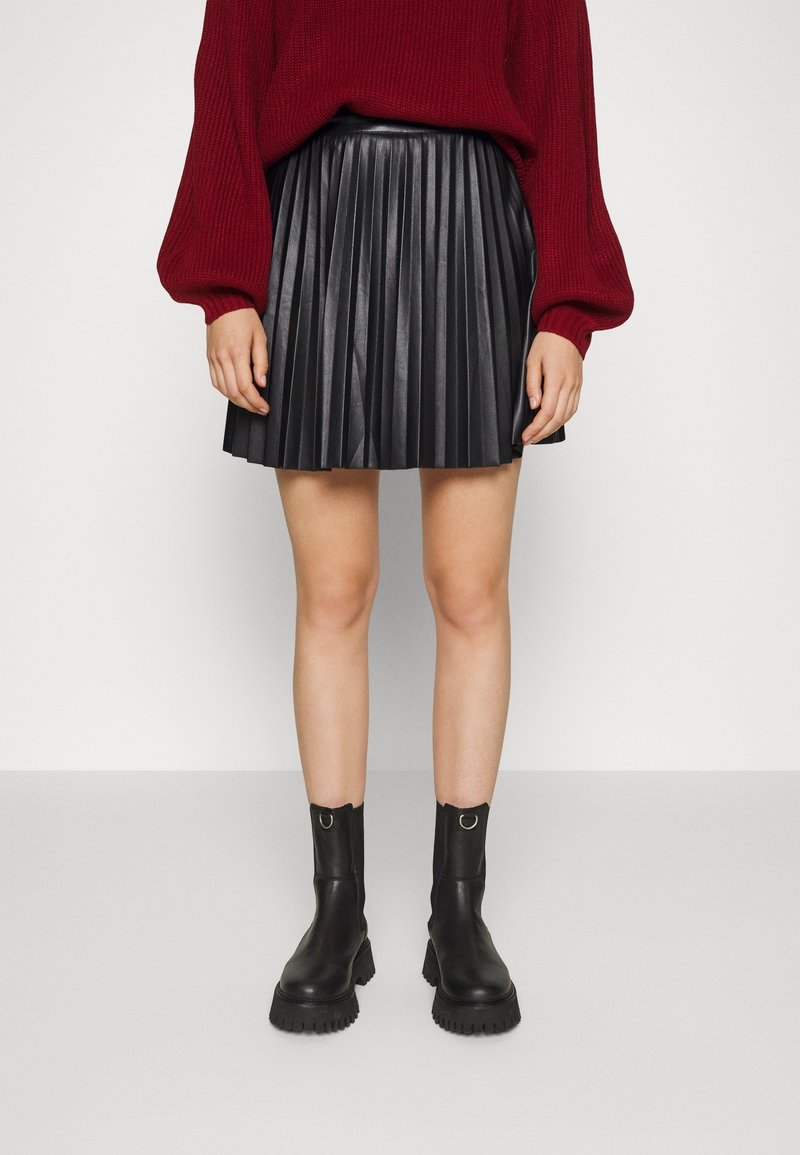 Femme portant un pull rouge, une jupe en cuir plissée noire et des bottines noires épaisses, debout devant un fond clair uni.