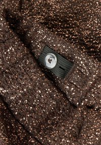 Tissu brun texturé avec des paillettes métalliques, doté d'un bouton visible. Le matériau semble tissé et scintillant, créant un aspect dimensionnel.