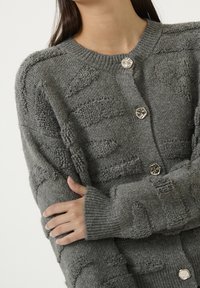 Cardigan grigio in maglia con motivi testurizzati, collo rotondo e bottoni in argento. Maniche lunghe con polsini a costine. Morbida texture chunky.