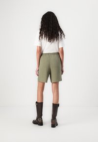 Olijfgroene shorts met een elastische tailleband, gecombineerd met bruin leren laarzen tot de knie. De achtergrond is volledig wit zonder verdere details.