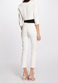 Kvinna som vänder sig bort iförd vit cropped kavaj, högmidjade vita byxor, svart topp och silverfärgade högklackade sandaler mot enfärgad bakgrund.