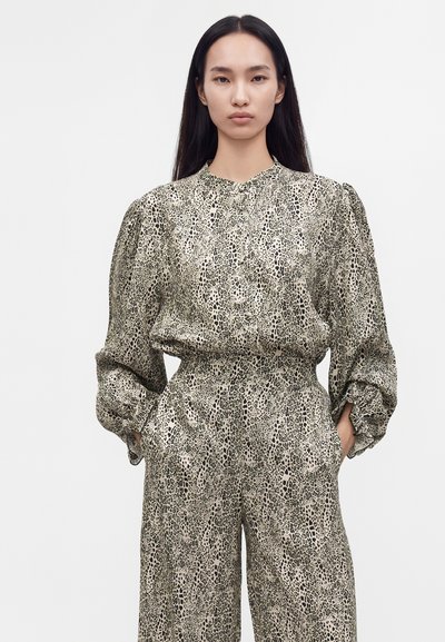 Femme aux longs cheveux foncés portant un haut à manches bouffantes et col montant avec un pantalon large assorti beige et noir à imprimé animal, mains dans les poches.