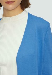 Blauwe cardigan van glad gebreid stof, gedragen over een witte geribde coltrui. Heeft een ontspannen pasvorm en een open voorkant.
