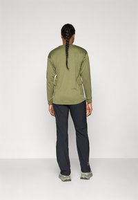 Olive green long-sleeve shirt with a relaxed fit, paired with black pants. Material appears lightweight, with side pockets and long sleeves.