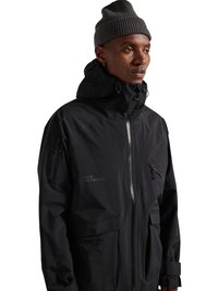 Mann trägt eine schwarze Kapuzenjacke mit Reißverschluss- und Klappentaschen und eine graue gestrickte Mütze und schaut nach rechts vor weißem Hintergrund.