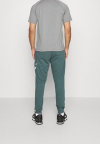 Hombre vestido con pantalones jogger en color teal con detalles reflectantes, camiseta gris de manga corta, calcetines blancos y zapatillas deportivas negras, de pie contra una pared blanca.