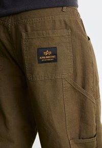 Olivengrønne cargo-bukser med en tekstureret finish, udstyret med et sort label-patch broderet med "ALPHA INDUSTRIES" og et orange logo.