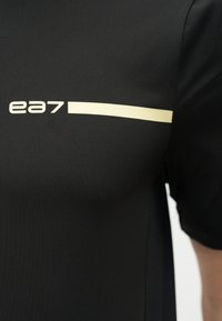 Μαύρο ύφασμα με κοντό μανίκι που εμφανίζει το λογότυπο "ea7" και μια οριζόντια κρεμ ρίγα στη περιοχή του στήθους ενός πουκαμίσου ή τοπ.