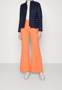 Granatowa pikowana kurtka puchowa ze stójką, zestawiona z jasnopomarańczowymi rozszerzanymi spodniami i jasnymi masywnymi sneakersami.