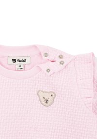 Pink cotton onesie featuring a textured herringbone pattern, ruffled accents on shoulders, and a bear embroidery on the chest. Snaps at the neckline.