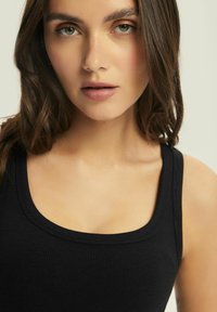 Czarny prążkowany top na ramiączkach z szerokimi paskami, charakteryzujący się okrągłym dekoltem i gładką teksturą. Zbliżenie na twarz, z delikatnymi rysami i naturalnym makijażem.