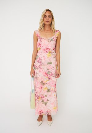 Kvinde i en pink blomsterprintet maxikjole uden ærmer med flæser, der holder en pastel-farvet perlet håndtaske og har hvide spidse hæle på.