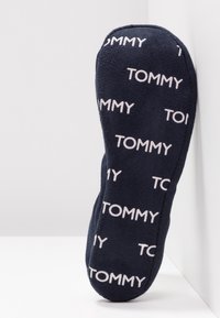 Semelle de chausson bleu marine avec un motif répétitif de texte "TOMMY" en blanc, placée debout contre un coin de mur blanc.