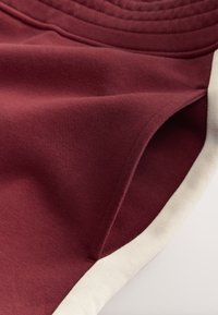 Bordeauxfarvet stof med en blød tekstur, der har en cremefarvet kant langs kanten og en subtil ribbet detalje øverst.