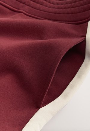 Bordeauxrode stof met een zachte textuur, voorzien van een crèmekleurige bies langs de rand en een subtiel geribd detail aan de bovenkant.