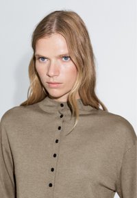 Beige långärmad topp med hög krage och åtta svarta knappar fram. Stickad struktur med en slät yta. Neutral färg.