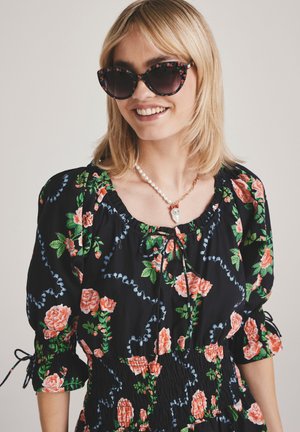 Vestido negro con flores y rosas rosas, con cintura fruncida, mangas abullonadas y detalles en azul. Los accesorios incluyen collares de perlas y cadenas doradas, y gafas de sol de gran tamaño.