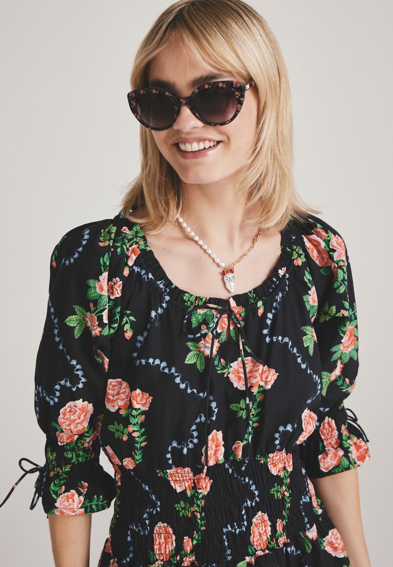 Rochie neagră cu flori și trandafiri roz, talie strânsă, mâneci bufante și detalii albastre. Accesoriile includ coliere din perlă și lanț de aur, ochelari de soare supradimensionați.