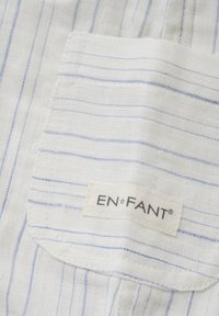 Weißer Stoff mit hellblauen Streifen, ausgestattet mit einer kleinen aufgesetzten Tasche und einem Etikett mit der Aufschrift "EN•FANT", aus einem strukturierten Material gefertigt.