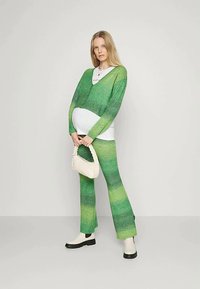 Mulher grávida vestindo um cardigã de malha em degradê verde e calças, camisa branca, segurando uma bolsa trançada de creme, de pé contra um fundo simples.