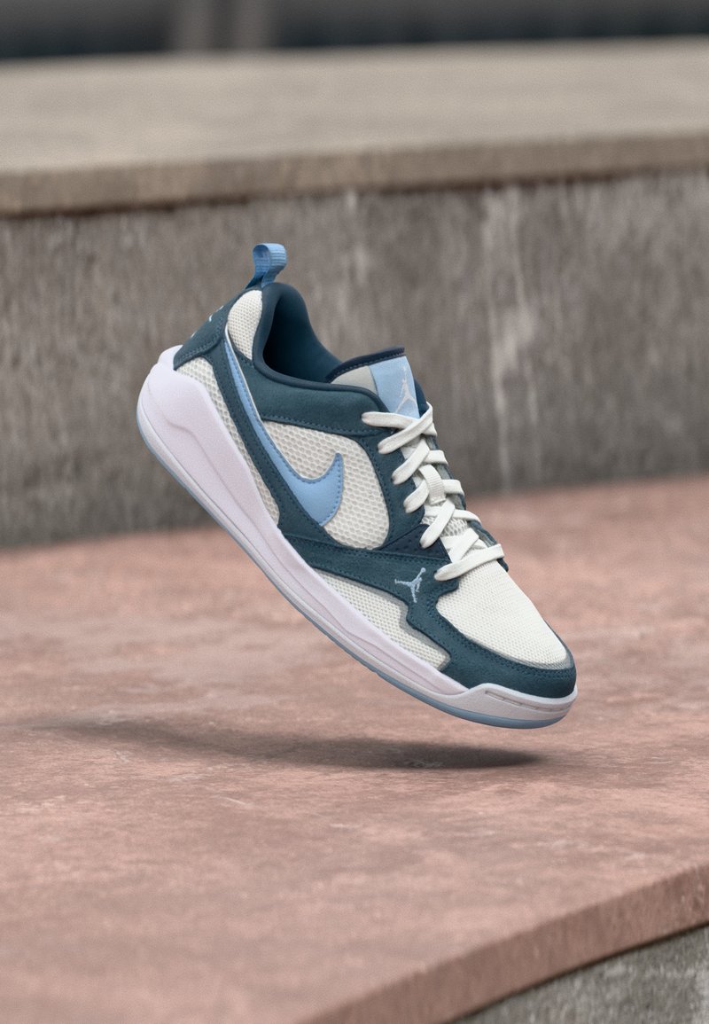 Baskets Nike Air Jordan bleues et blanches flottant au-dessus d'une surface en béton avec un fond en pierre flou.