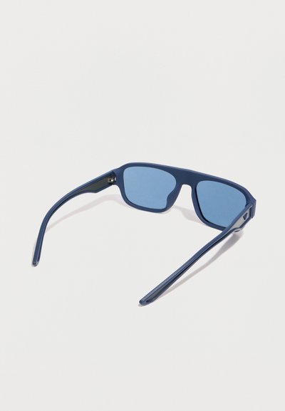 Μπλε ορθογώνια γυαλιά ηλίου με συμπαγείς μπλε φακούς και πλατιά καμπυλωτά πλαίσια, απεικονιζόμενα από πίσω γωνία σε λευκό φόντο.