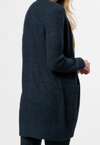 Long gilet bleu foncé en tissu tricoté texturé, avec des poignets côtelés et deux poches latérales, au design ample et ouvert.