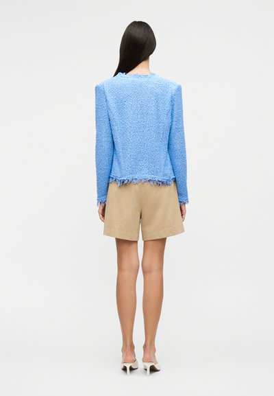 Γυναίκα που στέκεται γυρισμένη με την πλάτη, φοράει μπλε μπουφάν με υφή και μακριά μανίκια, μπεζ σορτς μήκους μέχρι το γόνατο και λευκά παπούτσια με τακούνι, σε λευκό φόντο.