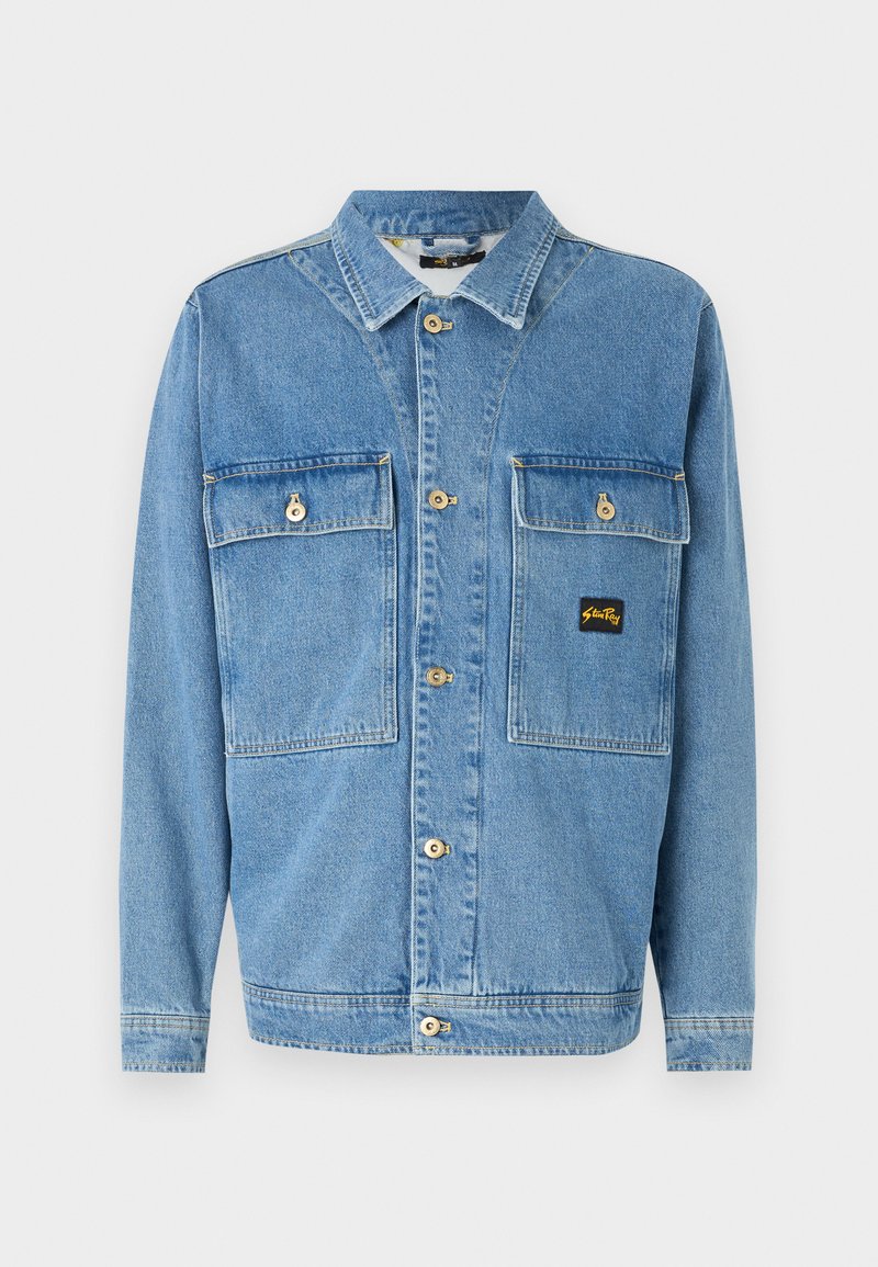 Veste en denim bleu clair avec un devant boutonné, deux poches poitrine avec rabats et un petit patch logo. Col classique et manches longues.
